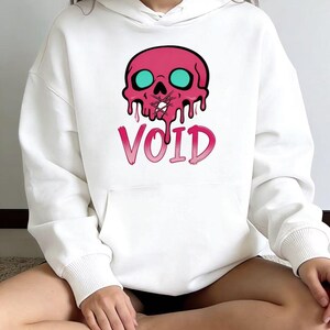 May include: White hoodie featuring a pink skull graphic with teal eyes and dripping details. The word "VOID" is printed below the skull in a gradient pink color. The hoodie has a front pocket and long sleeves.