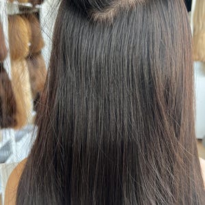 May include: A long, straight, dark brown hair extension piece attached to the top of the head. The hair is smooth and sleek, featuring a visible weft construction where the hair strands are secured at the base, creating a layered, full appearance.