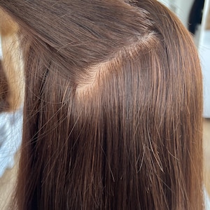 May include: A medium brown hairpiece featuring a visible monofilament base construction where individual strands are integrated into a sheer material, creating a natural-looking part line against the scalp with smooth, straight hair texture.
