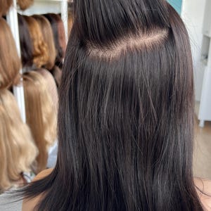 Puede incluir: Una peluca de color marrón oscuro con textura lisa y una raya de monofilamento visible en la coronilla. El cabello está peinado con una sección recogida en una coleta alta, revelando la base que imita el cuero cabelludo y mechones lisos.