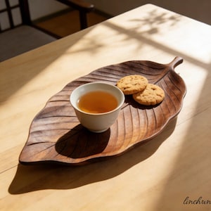 Puede incluir: Una bandeja de madera con forma de hoja con una taza de té blanca llena de té y dos galletas. La bandeja es de color marrón oscuro, con vetas talladas. El té es de color ámbar y las galletas son de color dorado. La escena está iluminada por luz natural.