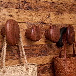 May include: Wooden wall hooks in various shapes and sizes, mounted on a wooden wall. The hooks are in shades of brown and are holding bags with rope and leather straps. The bags are made of woven materials.