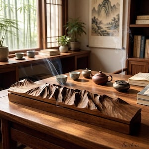 Peut inclure: Un porte-encens en bois sculpté avec un motif de chaîne de montagnes. Le support marron est posé sur une table en bois avec des tasses à thé, une théière et un brûleur d'encens. La fumée s'élève de l'encens, créant une atmosphère sereine.