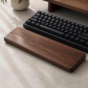 May include: A rectangular wooden wrist rest with a smooth, dark brown grain finish positioned in front of a black mechanical keyboard. The wrist rest features rounded edges and a solid construction, designed to sit parallel to the keyboard.