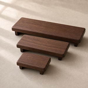 Peut inclure: Trois rehausseurs rectangulaires en bois brun foncé de longueurs variables, chacun doté d'une surface plane aux bords arrondis et de quatre pieds courts et robustes. Les rehausseurs sont disposés en cascade, du plus grand au plus petit.