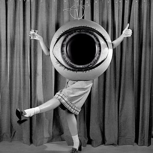 May include: A person wearing a large, inflatable eye costume with a black pupil and white sclera. The person is holding a glass in one hand and giving a thumbs up with the other. The person is standing in front of a curtain.