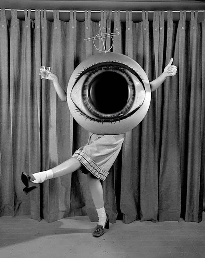 May include: A person wearing a large, inflatable eye costume with a black pupil and white sclera. The person is holding a glass in one hand and giving a thumbs up with the other. The person is standing in front of a curtain.