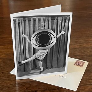 May include: A black and white photograph of a person wearing a giant eye costume. The person is standing in front of a curtain and is holding a glass in one hand and a thumbs up in the other.