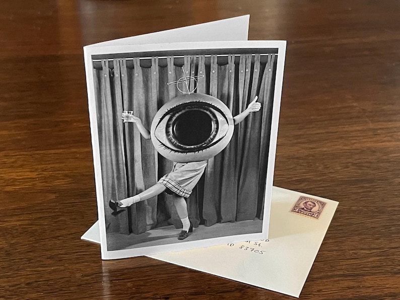 May include: A black and white photograph of a person wearing a giant eye costume. The person is standing in front of a curtain and is holding a glass in one hand and a thumbs up in the other.