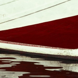 May include: A small, white canoe with red trim floats on the water. The canoe is positioned in front of a larger boat with a red hull and white upper section. A rope is tied to the canoe's bow. The water reflects the boat and canoe.