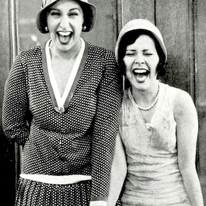 May include: A monochrome image of two women laughing. One is in a polka dot dress and a cloche hat, the other in a sleeveless dress and a pearl necklace. They are holding hands, evoking a vintage style.