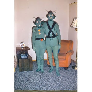 May include: A couple dressed in green alien costumes with antennas and masks. They are standing in a living room with a brown end table and a lamp.