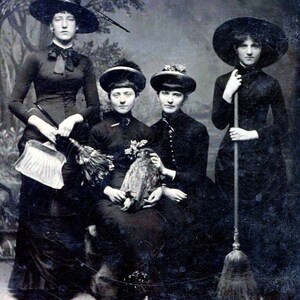 May include: Four women dressed in black clothing and hats pose for a photo. Three of the women are wearing witch hats, and one is holding a broom. The woman in the center is holding a stuffed owl.