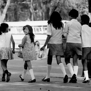 May include: Four children, three girls and one boy, walking along a pavement. The girls are wearing dresses and the boy is wearing shorts. They are all smiling and looking happy.