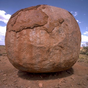 May include: A large, round, red rock with a rough, textured surface sits on a red dirt ground. The rock is in the foreground of the image, with a blue sky and white clouds in the background.