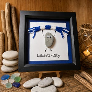 May include: A framed pebble art piece featuring a grey stone with googly eyes, wearing a blue and white crocheted scarf. The stone has drawn arms and legs, and the text Leicester City is printed below on a white background with a blue border.