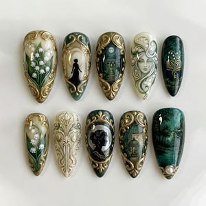 May include: A set of ten almond-shaped press-on nails with intricate designs. The nails feature a variety of art, including floral patterns, silhouette figures, and landscape scenes, all framed with gold accents. The color palette includes green, white, and gold.