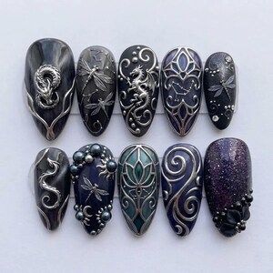 May include: A set of ten press-on nails featuring dark purple and black bases with intricate silver metallic embellishments, including dragon figures, dragonfly motifs, celestial patterns, floral shapes, and small dark beads in various sizes.