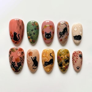 Peut inclure: Un ensemble de dix faux ongles décorés sur le thème de l'automne avec des chats noirs, des feuilles mortes, des glands et des motifs floraux. Les ongles présentent diverses finitions dont des paillettes, des textures brillantes et des gouttes d'eau.