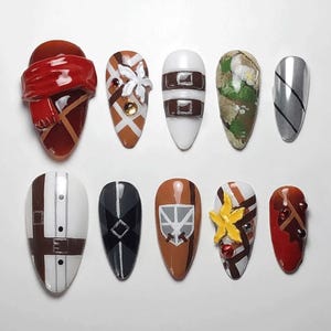 May include: A collection of ten almond-shaped fingernails, each painted with a unique design. The designs include a red scarf, white flower, brown straps, camouflage, silver stripes, a white shirt, a black shield, a brown shield, a yellow star, and red gems.