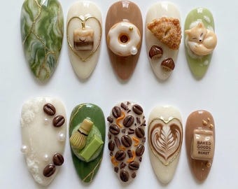 May include: A set of ten almond-shaped artificial nails with various designs. The designs include coffee beans, a donut, a sleeping fox, a latte art heart, and a small jar with the text "Baked Goods Roast."