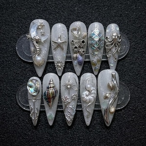 May include: A set of ten long, almond-shaped artificial nails. Each nail is painted with a shimmering, glittery white polish and adorned with intricate, silver-toned sea-themed embellishments, including seahorses, starfish, and shells.