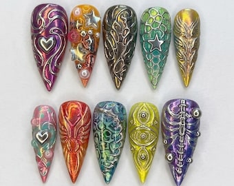 May include: A set of ten almond-shaped press-on nails featuring diverse 3D designs. The collection includes metallic purple with a heart, orange with stars and pearls, dark textured patterns, green scales with a star, gold wings, pink with a heart, red flame-like textures, blue marbled patterns, yellow with eye motifs, and purple with a skeletal rib cage design.