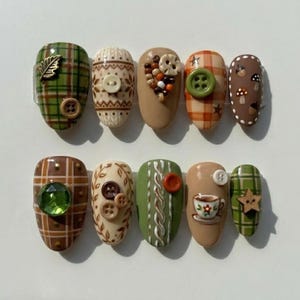 May include: A set of ten press-on nails featuring autumn-themed designs including green and brown plaid patterns, cable knit textures, miniature buttons, a small teacup illustration, tiny mushrooms, acorns, a gold leaf charm, and a green gem.