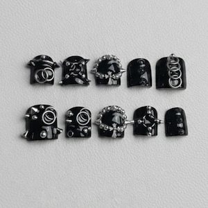 Può includere: Unghie finte nere con vari abbellimenti argentati. I disegni includono anelli d'argento, borchie ed elementi decorativi. Le unghie sono lucide e hanno un'estetica scura e gotica.