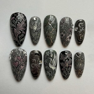 Könnte beinhalten: Zehn künstliche Fingernägel mit aufwendigen Designs. Die Nägel zeigen verschiedene Muster, darunter Blumen-, Vogel- und Schmetterlingsmotive, in Schwarz-, Grau- und Rosatönen. Einige Nägel sind mit Strasssteinen verziert.