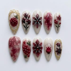 May include: A collection of ten almond-shaped false nails. Each nail showcases a distinct design, such as red flowers, berries, and water droplet effects. Some nails have gold accents and small bee illustrations. The color scheme includes red, white, and gold hues.