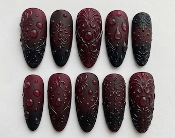 May include: A set of ten almond-shaped press-on nails in a deep burgundy and black gradient. Each nail features raised, glossy, three-dimensional filigree scrollwork and small, clear, rounded droplet accents against a matte base finish.