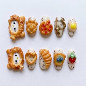 May include: A set of ten artificial nails with various food-themed designs. The designs include a lion, pizza, croissant, fried egg, toast with blueberries, and a strawberry. The nails are painted with a neutral base color and detailed with miniature food art.