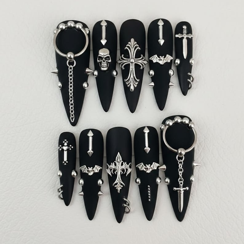May include: Black matte acrylic nails with silver embellishments. Designs include a skull, crosses, bats, and swords. Some nails feature chains, spikes, and rings for a gothic aesthetic. The nails are long and pointed.