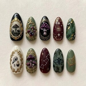 May include: A set of ten almond-shaped press-on nails featuring intricate designs including mushrooms, gold filigree frames, a skull, a potion bottle, constellations, a moth, and an open book, rendered in a palette of black, olive green, cream, and deep burgundy.