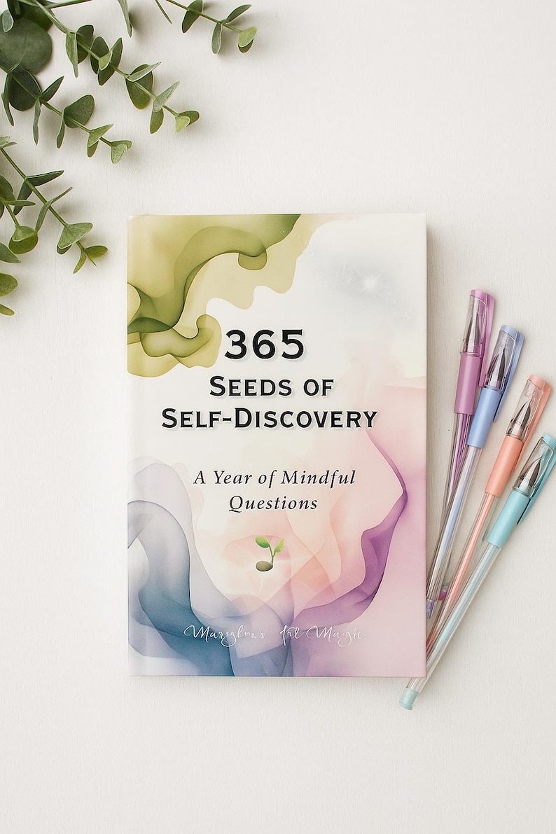 May include: A book titled "365 Seeds of Self-Discovery" with the subtitle "A Year of Mindful Questions." The cover features watercolor-style art in shades of green, pink, and purple. Several pastel-colored pens are arranged beside the book.