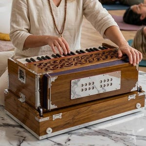 May include: A woman playing a wooden harmonium with a decorative top. The instrument is brown with silver accents. Other people are lying down on mats in the background. There are gongs and crystal bowls in the room.
