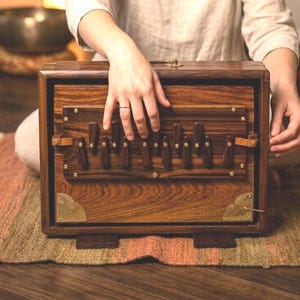 Op de afbeelding: Een houten harmonium met messing details. Het instrument is donkerbruin van kleur en heeft een reeks houten toetsen. Het harmonium staat op een kleurrijk tapijt. De afbeelding toont een persoon die het harmonium bespeelt.