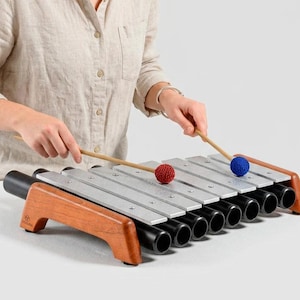 May include: A xylophone with silver metal bars and black cylindrical resonators set in a wooden frame. Two wooden mallets with textured red and blue spherical heads are positioned above the bars to produce musical notes.