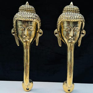May include: Two golden Buddha-head door handles. Each handle features a detailed depiction of Buddha's face, with closed eyes and a serene expression. The handles are elongated, with a rounded base for mounting. The background is black and white.