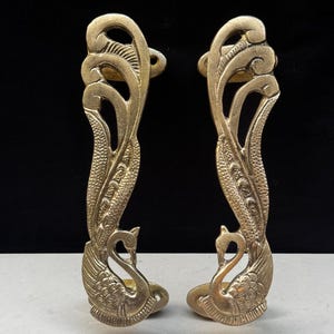 May include: Two brass-colored, ornate swan-shaped drawer pulls. Each pull features a swan at the base with a stylized feather design extending upwards. The pulls are designed with a loop at the top for mounting.