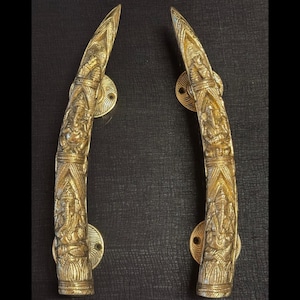 Op de afbeelding: Twee sierlijke, goudkleurige deurgrepen. Elke handgreep is gebogen en heeft gedetailleerde gravures van figuren en patronen. De handgrepen zijn gemonteerd op ronde, decoratieve achterplaten.