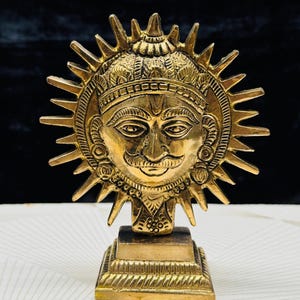 May include: A detailed brass sculpture of a sun face with a mustache and crown, surrounded by sun rays. The sculpture is mounted on a square base. The piece has an antique finish and is likely a decorative item.
