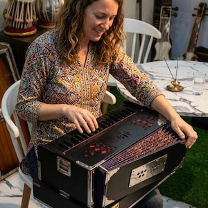 May include: A black harmonium musical instrument featuring a keyboard, a bellows system with purple patterned fabric, and decorative cutouts on the top panel. The instrument has metallic corner reinforcements and a rectangular metal plate with holes.