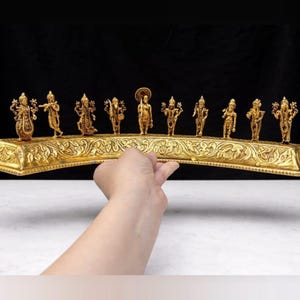 May include: A gold-coloured decorative piece with small, detailed figurines on an ornate, curved base. The figurines are likely religious figures. The base is held by a hand against a black backdrop.