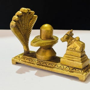 May include: A brass religious sculpture featuring a central lingam, a cobra hood, and a small bull figure. The sculpture is mounted on a rectangular base with ornate detailing. The overall colour is a warm, antique gold.