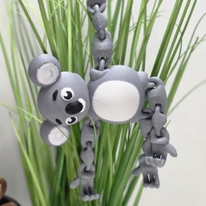 May include: A grey articulated koala bear toy with a white belly, ears, and eyes. The koala is hanging in front of green grass. The toy has a chain-like design for its limbs, allowing for flexible posing.