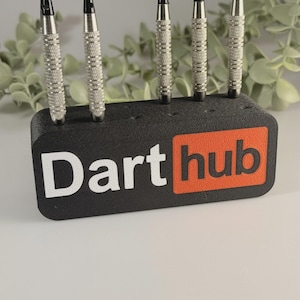 May include: A black rectangular dart stand with the text Dart hub printed on the front in white and orange. The stand features five circular holes along the top edge, with four silver and black metal darts inserted into the stand in an upright position.