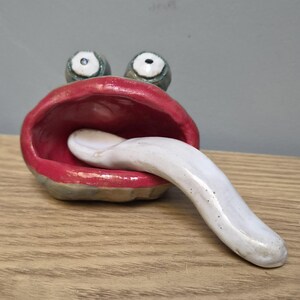 May include: A ceramic sculpture of a creature with a wide, open mouth featuring bright red lips and a long, protruding white tongue. The figure has two prominent, round eyes with white irises and dark pupils positioned above the mouth.