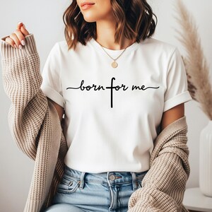Puede incluir: Camiseta blanca con la frase "born for me" en fuente negra, con una cruz reemplazando la letra "f". La camiseta se combina con vaqueros azules y una rebeca beige. Se ve un collar dorado.